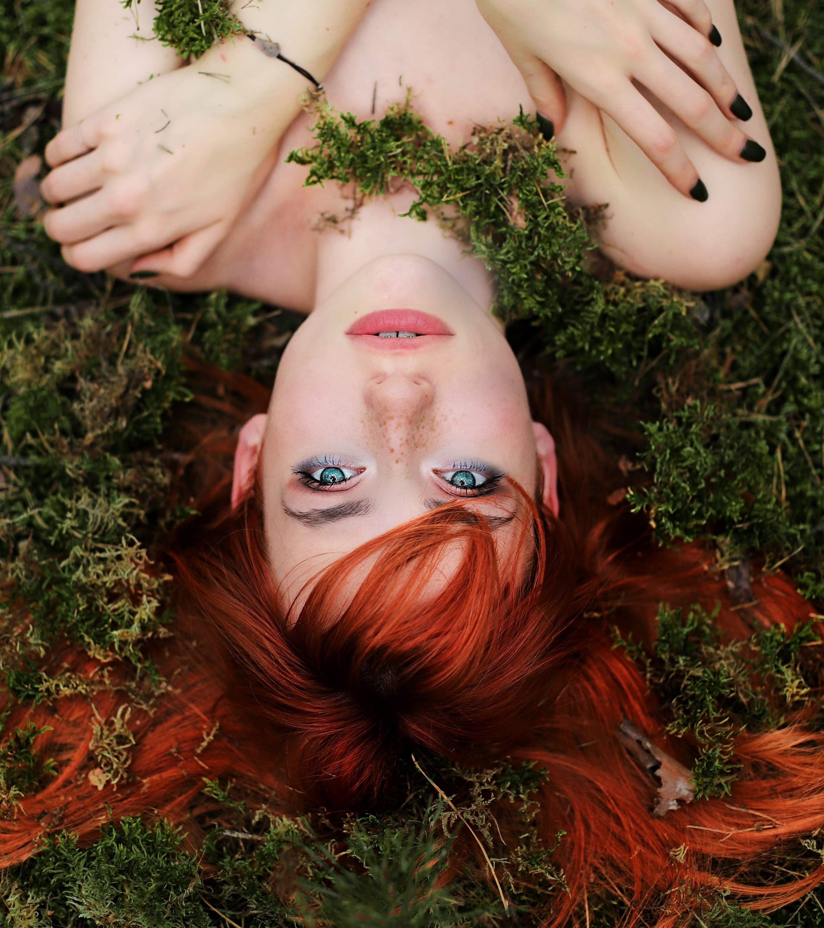 Portret rudej kobiety leżącej na mchu, z zielonym mchem wokół szyi i na włosach, patrzącej prosto w obiektyw swoimi intensywnie niebieskimi oczami, z ciemnym makijażem i czarnym manicure.