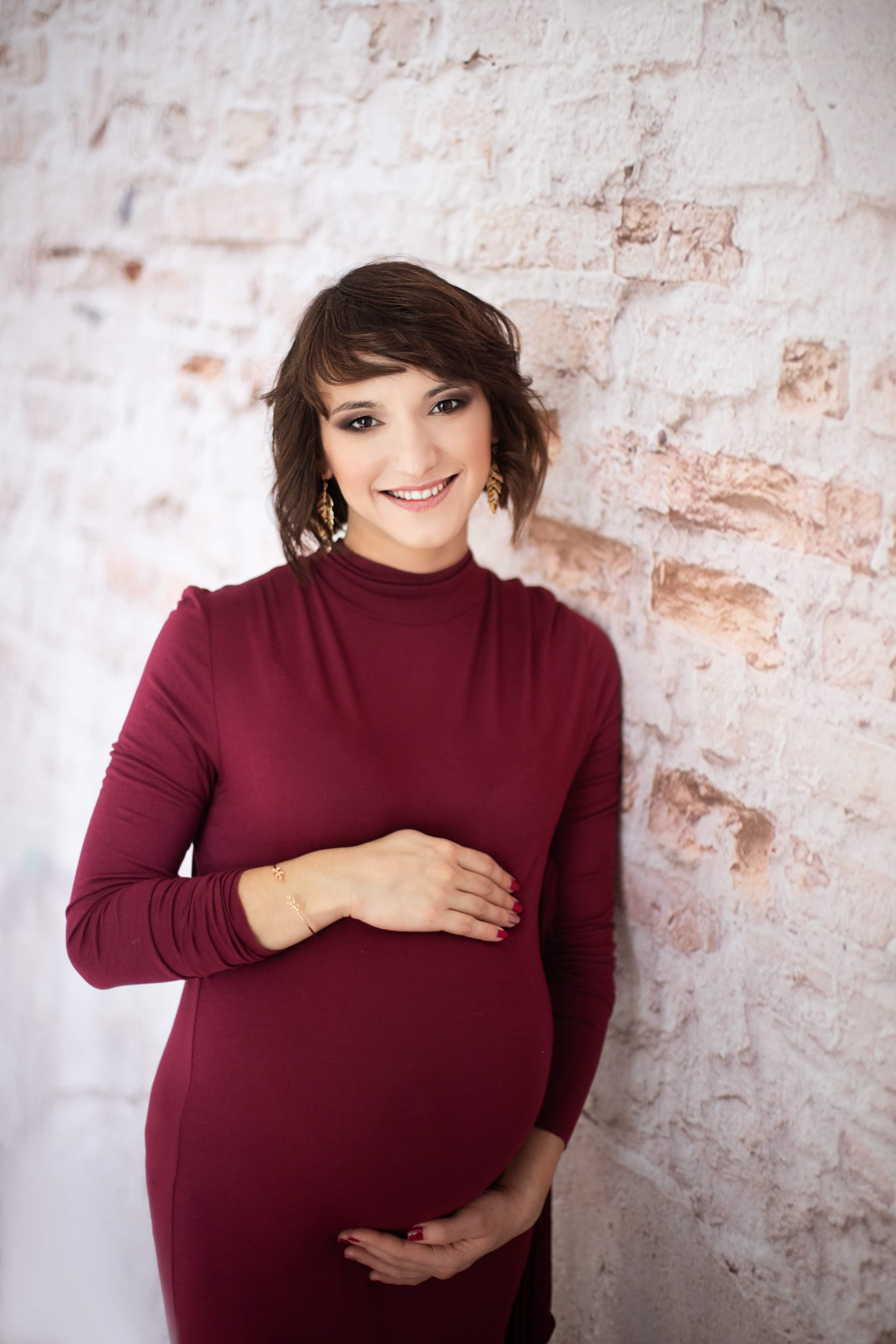 Uśmiechnięta kobieta w bordowej sukience ciążowej, pozująca na tle ściany z cegieł w odcieniach bieli i beżu, z delikatną biżuterią.