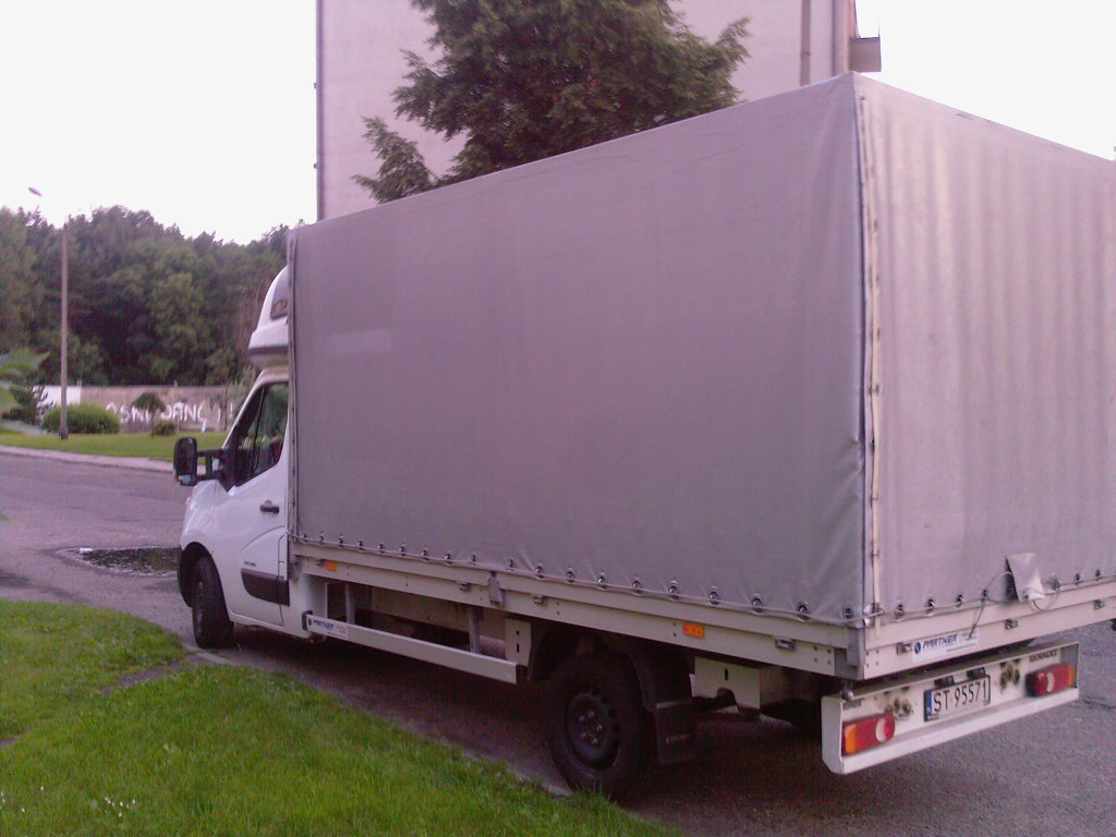 Biały samochód dostawczy z plandeką zaparkowany na poboczu, widok z boku, tablica rejestracyjna widoczna.