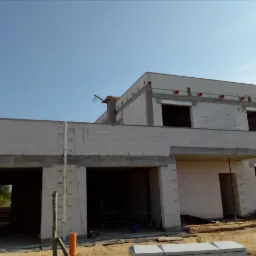 Surowy stan dwukondygnacyjnego domu z betonu komórkowego w trakcie budowy, widoczne otwarte garaże, okna bez ram i strop z wystającymi rurami wentylacyjnymi.