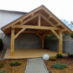 Drewniana altana z prostym dachem dwuspadowym, podłogą z desek i kamienną ścieżką prowadzącą do wejścia, otoczona zielenią i ogrodzeniem.