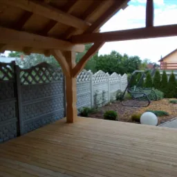 Zadaszony taras z drewnianą podłogą, widok na ogród z huśtawką, ogrodzenie z betonowych przęseł i kuliste lampy ogrodowe.