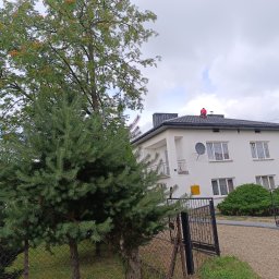 Bogdanexbud - Pracownik w czerwonej koszulce siedzi na ciemnoszarym dachu dwupiętrowego, białego domu z widoczną anteną satelitarną i ozdobnym drzewem jarzębiny z pomarańczowymi owocami na tle pochmurnego nieba.