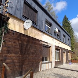 Bogdanexbud - Budynek w trakcie wymiany dachu na blachę trapezową w kolorze grafitowym, widoczne rusztowanie, instalacje elektryczne i satelitarna oraz fragment drewnianej elewacji i tarasu.
