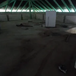 Poddasze z zieloną więźbą dachową i oknami w tle. Widoczne materiały budowlane i elementy konstrukcyjne. Wnętrze w trakcie prac, prawdopodobnie ocieplania.