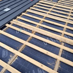 Bogdanexbud - Fragment dachu w trakcie krycia: szara blacha trapezowa ułożona częściowo, widoczna drewniana konstrukcja łat i kontrłat na membranie dachowej z nadrukiem.