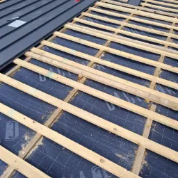 Fragment dachu w trakcie krycia: szara blacha trapezowa ułożona częściowo, widoczna drewniana konstrukcja łat i kontrłat na membranie dachowej z nadrukiem.