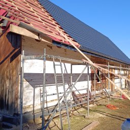 Bogdanexbud - Częściowo pokryty, ciemny dach z blachy trapezowej na drewnianej konstrukcji domu. Widoczne rusztowania i elementy konstrukcyjne dachu. Prace dekarskie w toku.
