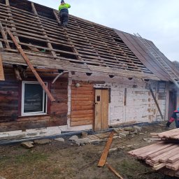 Bogdanexbud - Demontaż starego dachu drewnianego domu. Widoczna więźba dachowa i częściowo zdjęte pokrycie. Na dachu pracuje dekarz w jaskrawej kamizelce. Przygotowane nowe deski.