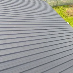 Szary dach z blachy trapezowej, widok z góry. W tle zieleń i fragment szklarni.