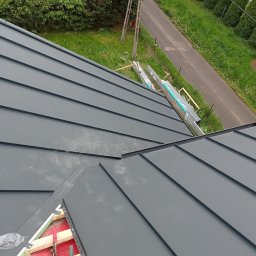 Bogdanexbud - Montaż blachy na dachu. Widoczne narzędzia dekarskie, elementy konstrukcji i blacha w kolorze grafitowym. W tle zieleń i droga.