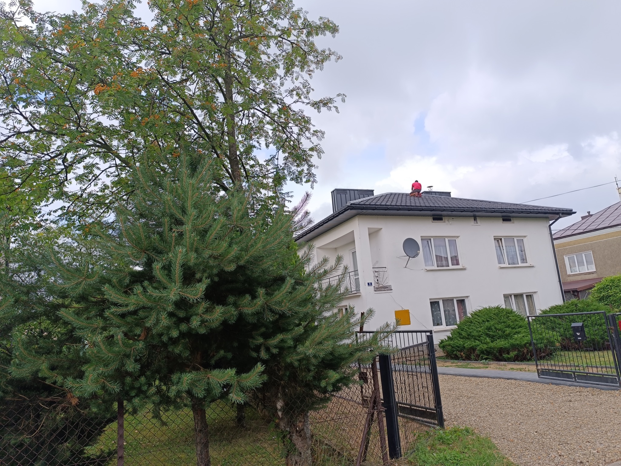 Pracownik w czerwonej koszulce siedzi na ciemnoszarym dachu dwupiętrowego, białego domu z widoczną anteną satelitarną i ozdobnym drzewem jarzębiny z pomarańczowymi owocami na tle pochmurnego nieba.