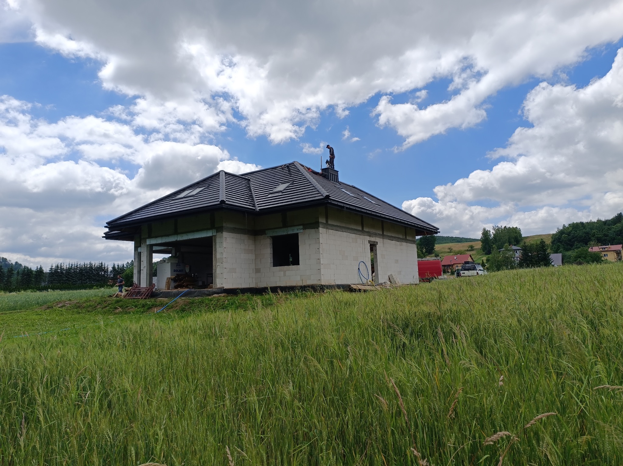 Dom w trakcie budowy z ułożoną dachówką, widoczny dekarz na kominie, otoczony wysoką trawą i polami uprawnymi, na tle błękitne niebo z chmurami.