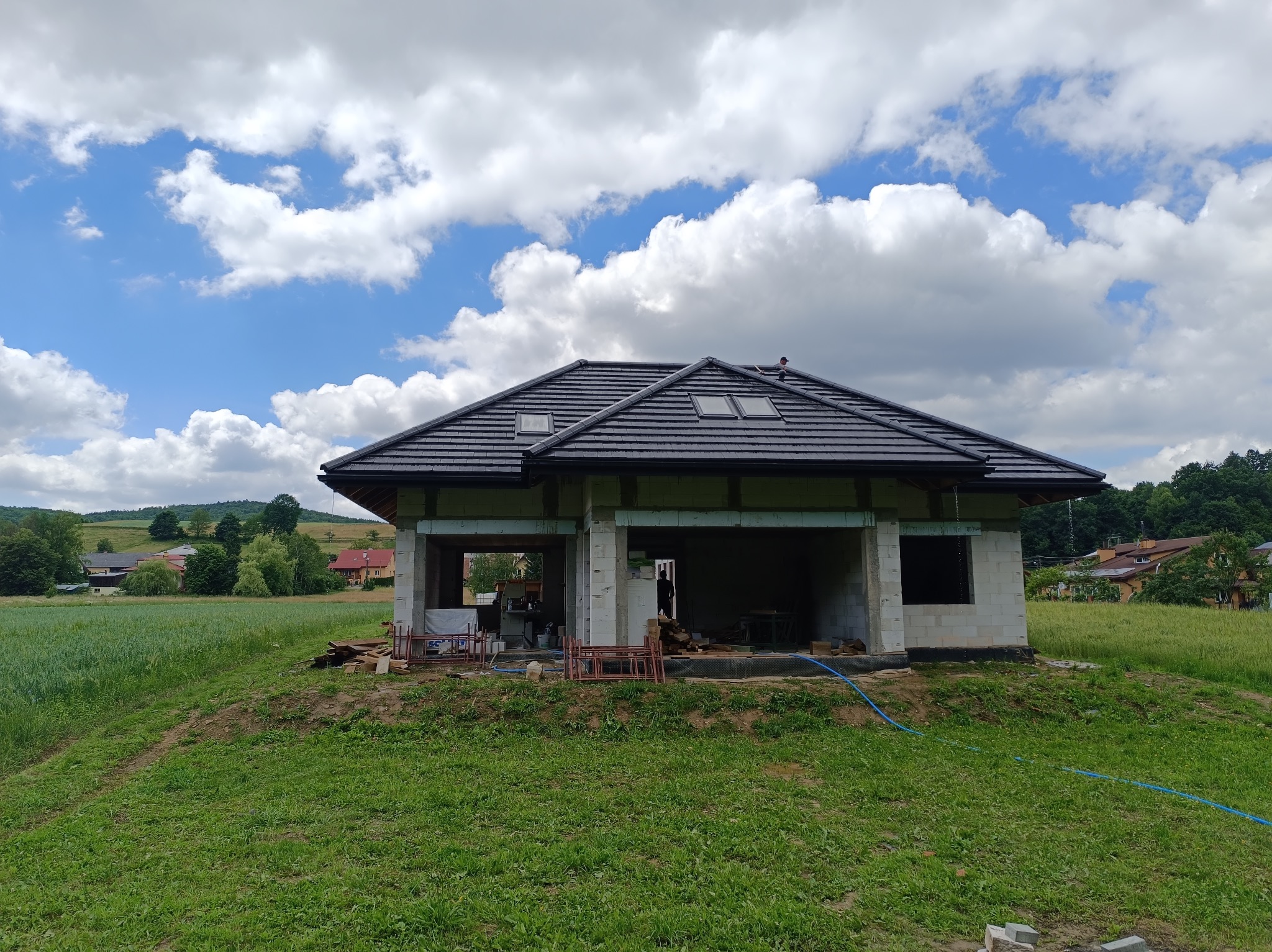 Surowy dom jednorodzinny w trakcie budowy, z widocznym nowym, czarnym dachem z dachówkami i oknami dachowymi, na tle zielonego krajobrazu i błękitnego nieba z chmurami.