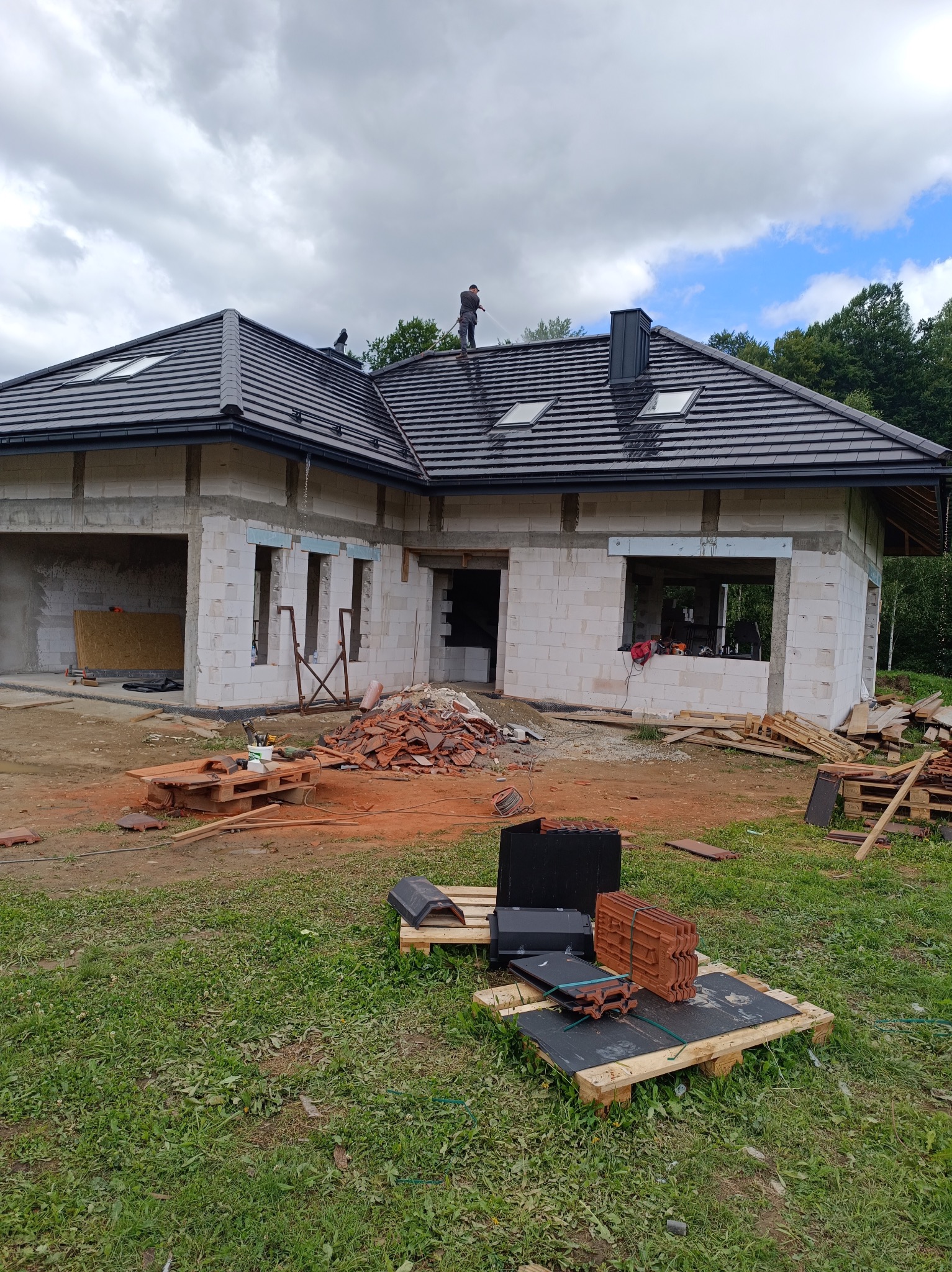 Budowa domu jednorodzinnego w trakcie, widoczny nowy dach z czarnej dachówki, dwóch dekarzy na dachu, nieotynkowane ściany z białego betonu komórkowego, palety z dachówkami na trawniku.