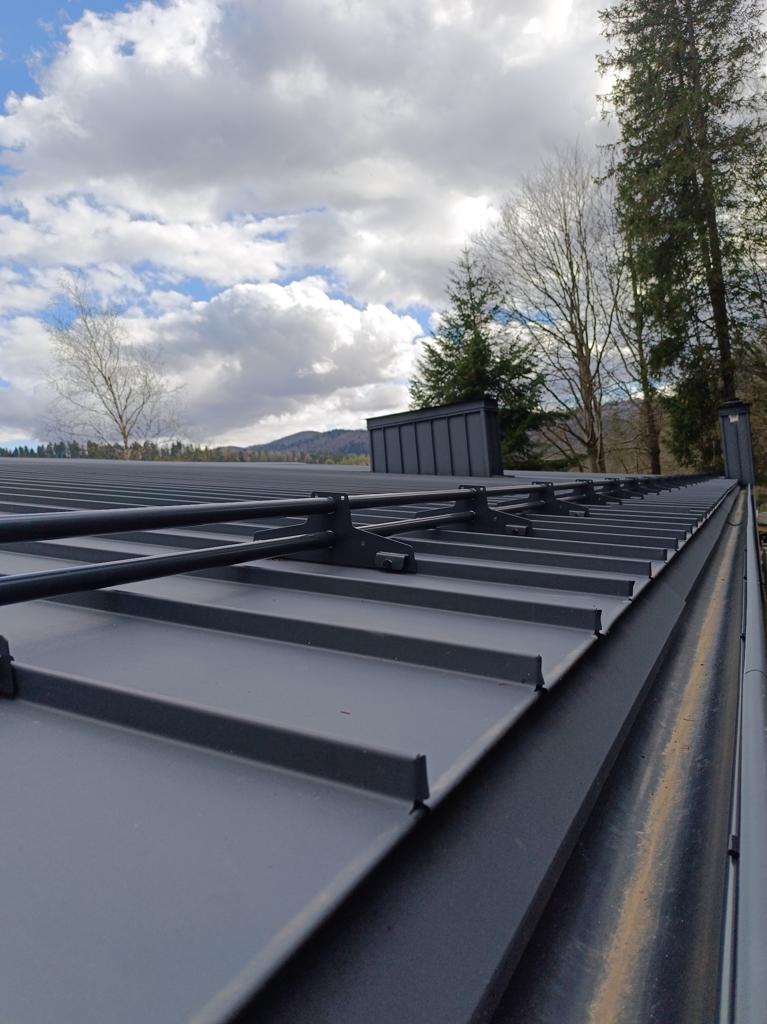 Szary dach z blachy trapezowej z zamontowanymi wspornikami i rurami odgromowymi na tle pochmurnego nieba i drzew