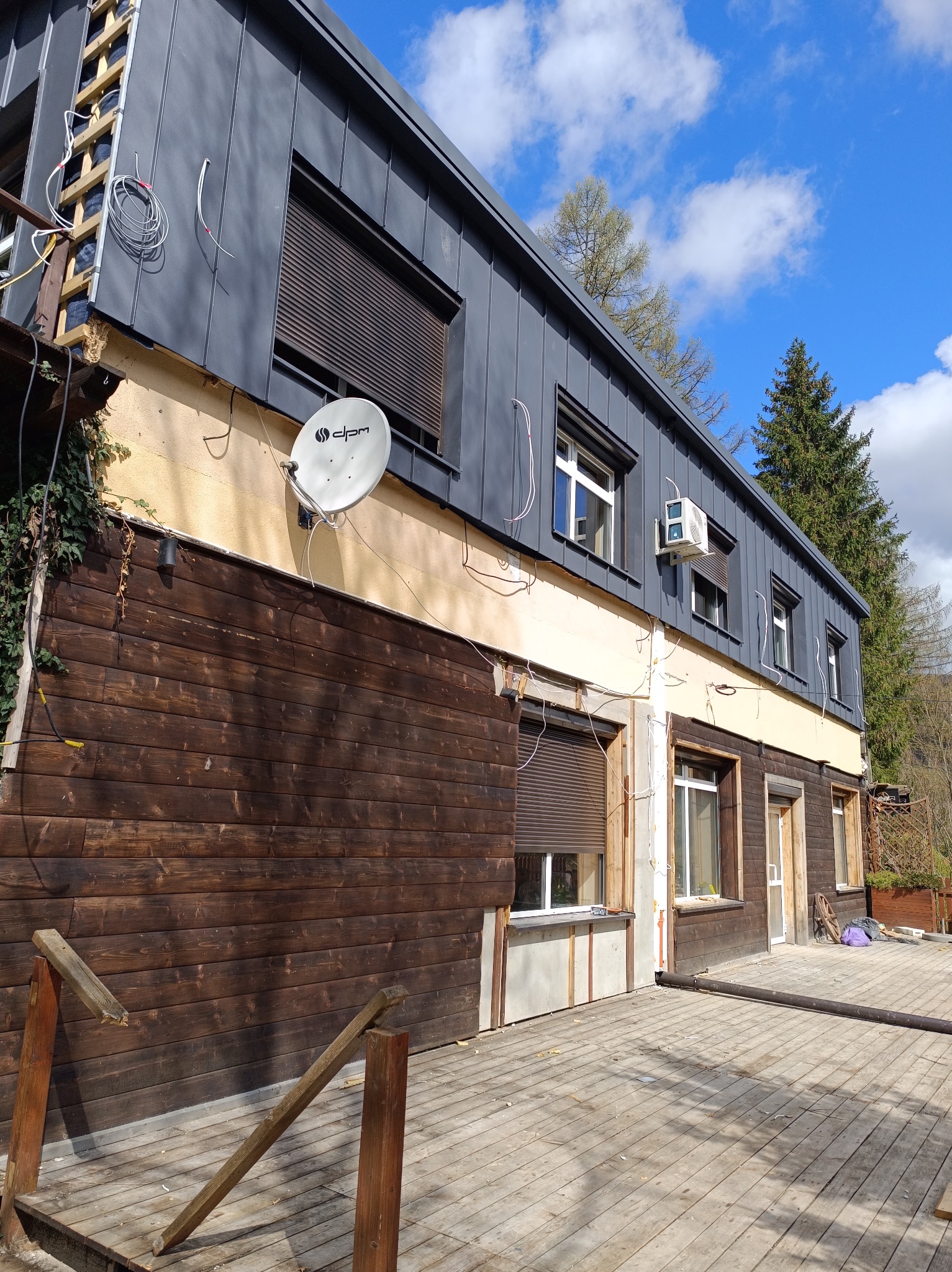 Budynek w trakcie wymiany dachu na blachę trapezową w kolorze grafitowym, widoczne rusztowanie, instalacje elektryczne i satelitarna oraz fragment drewnianej elewacji i tarasu.