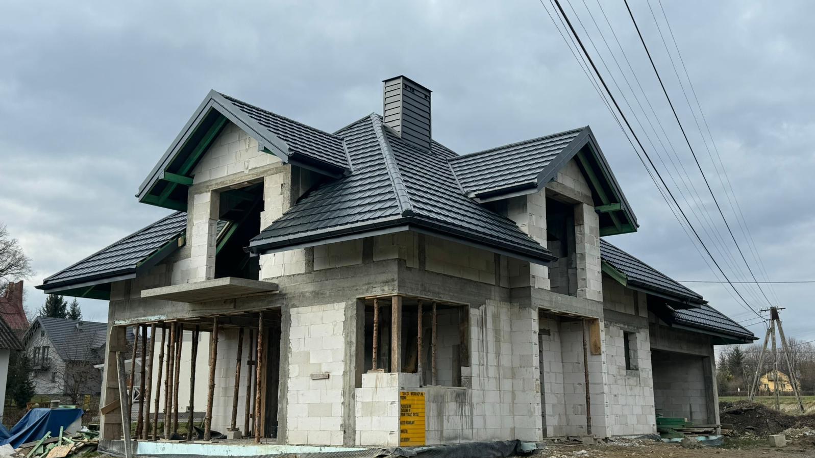 Dom w trakcie budowy z zamontowanym, czarnym dachem z blachodachówki, widoczna konstrukcja budynku z betonu komórkowego i drewniane podpory.