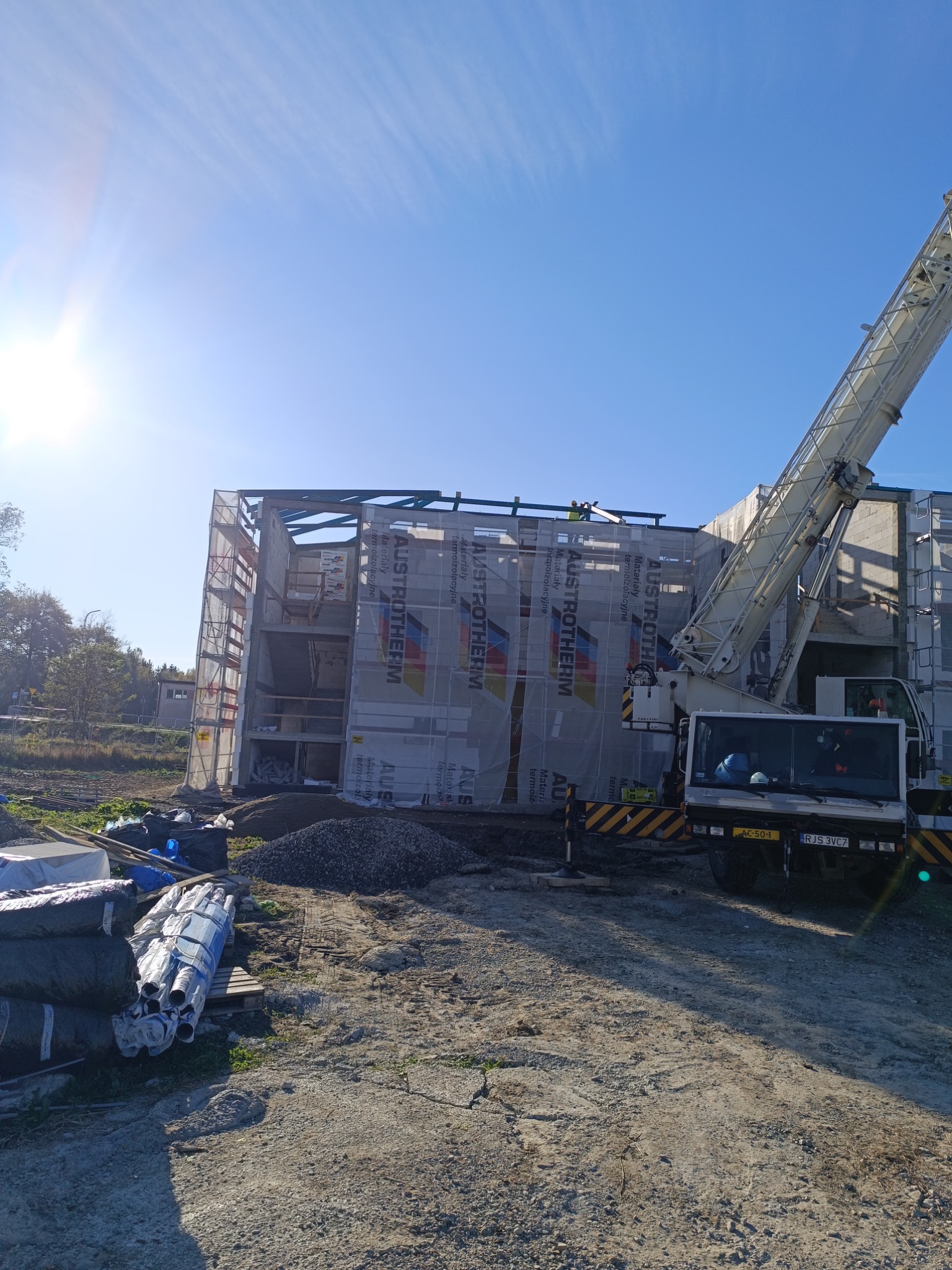 Budowa domu z widocznym rusztowaniem, dźwigiem i materiałami izolacyjnymi Austrotherm, częściowo osłonięta siatką ochronną.