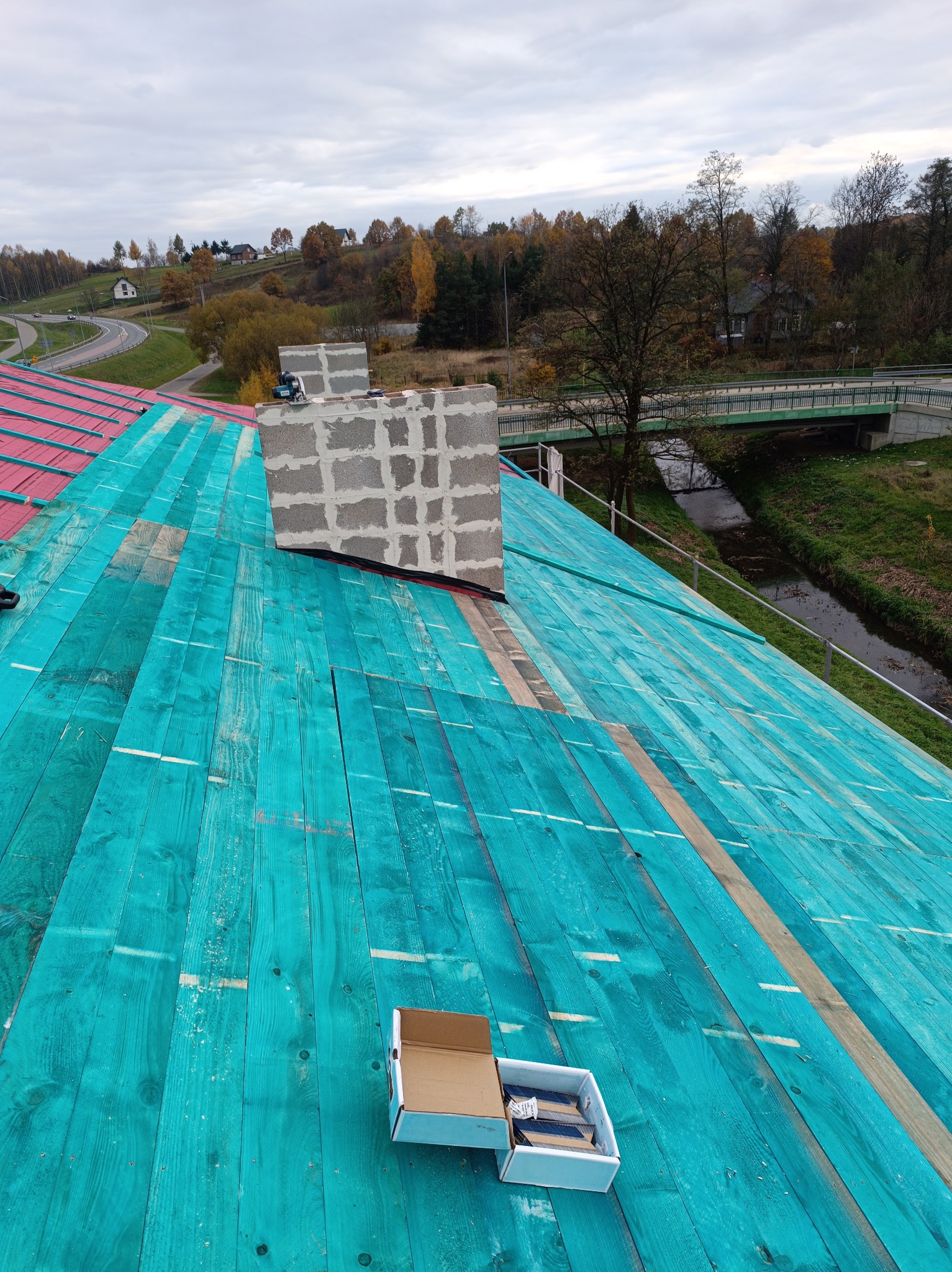 Widok z góry na konstrukcję dachu z desek pomalowanych na turkusowo, z widocznym kominem z szarych pustaków, pudełkiem gwoździ dekarskich i okolicznym krajobrazem z rzeką oraz drogą.
