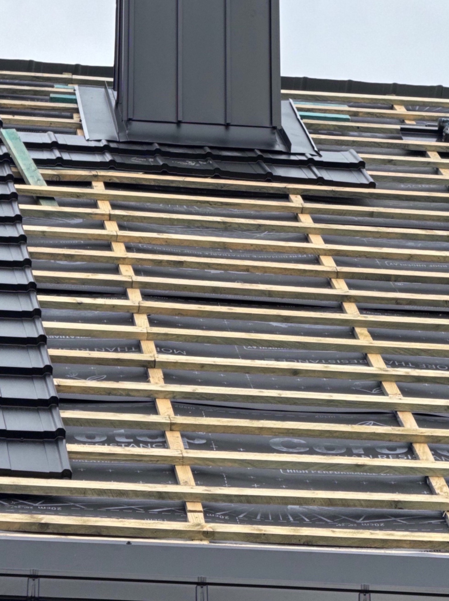 Czarny komin z blachy na dachu z częściowo ułożoną dachówką. Widoczne łaty i membrana dachowa. Montaż dachu na ukończonym etapie.