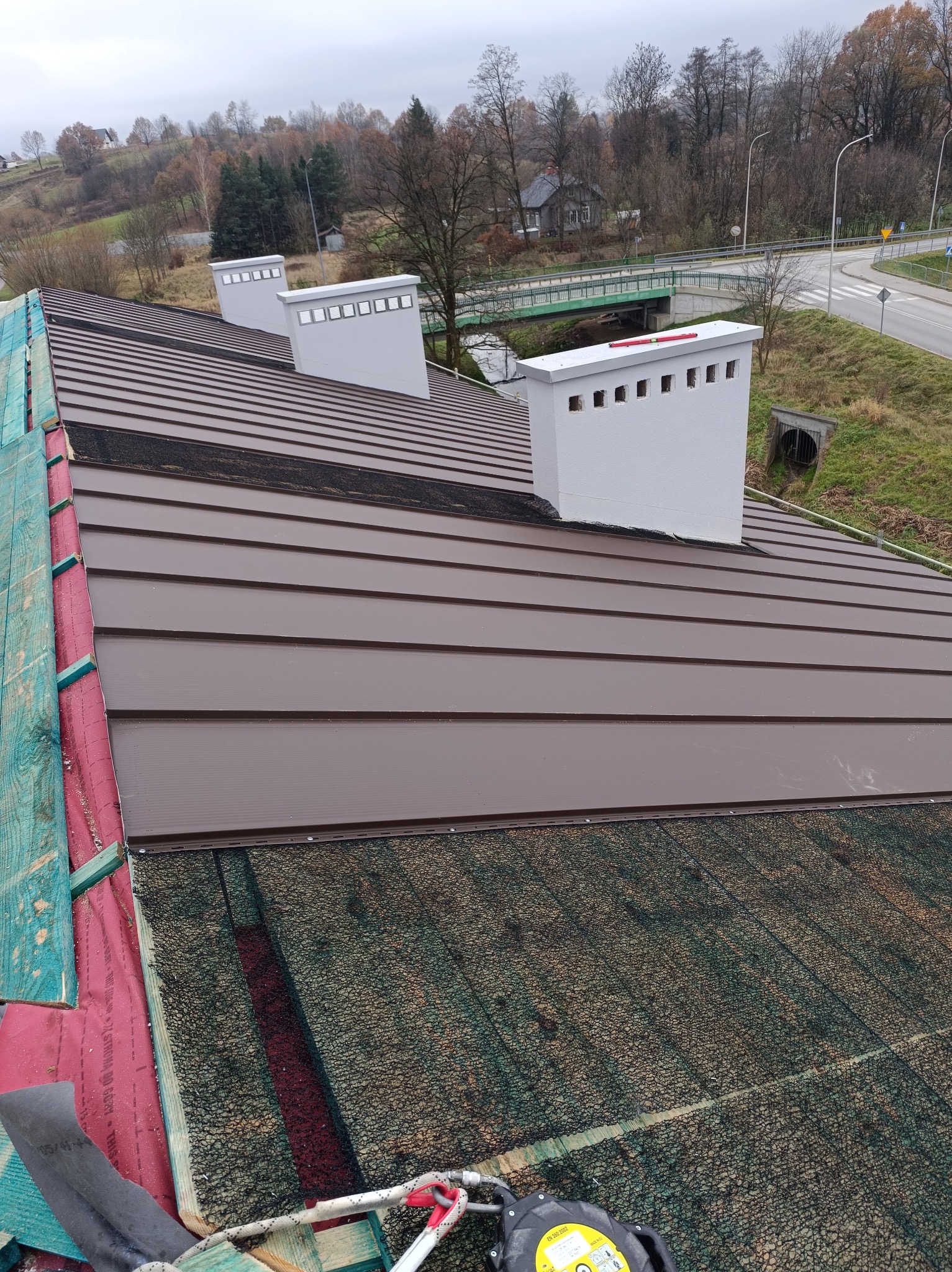 Brązowy dach z blachy na rąbek stojący, trzy białe kominy wentylacyjne z otworami, zabezpieczenia na dachu, widok na okolicę, dzień pochmurny.