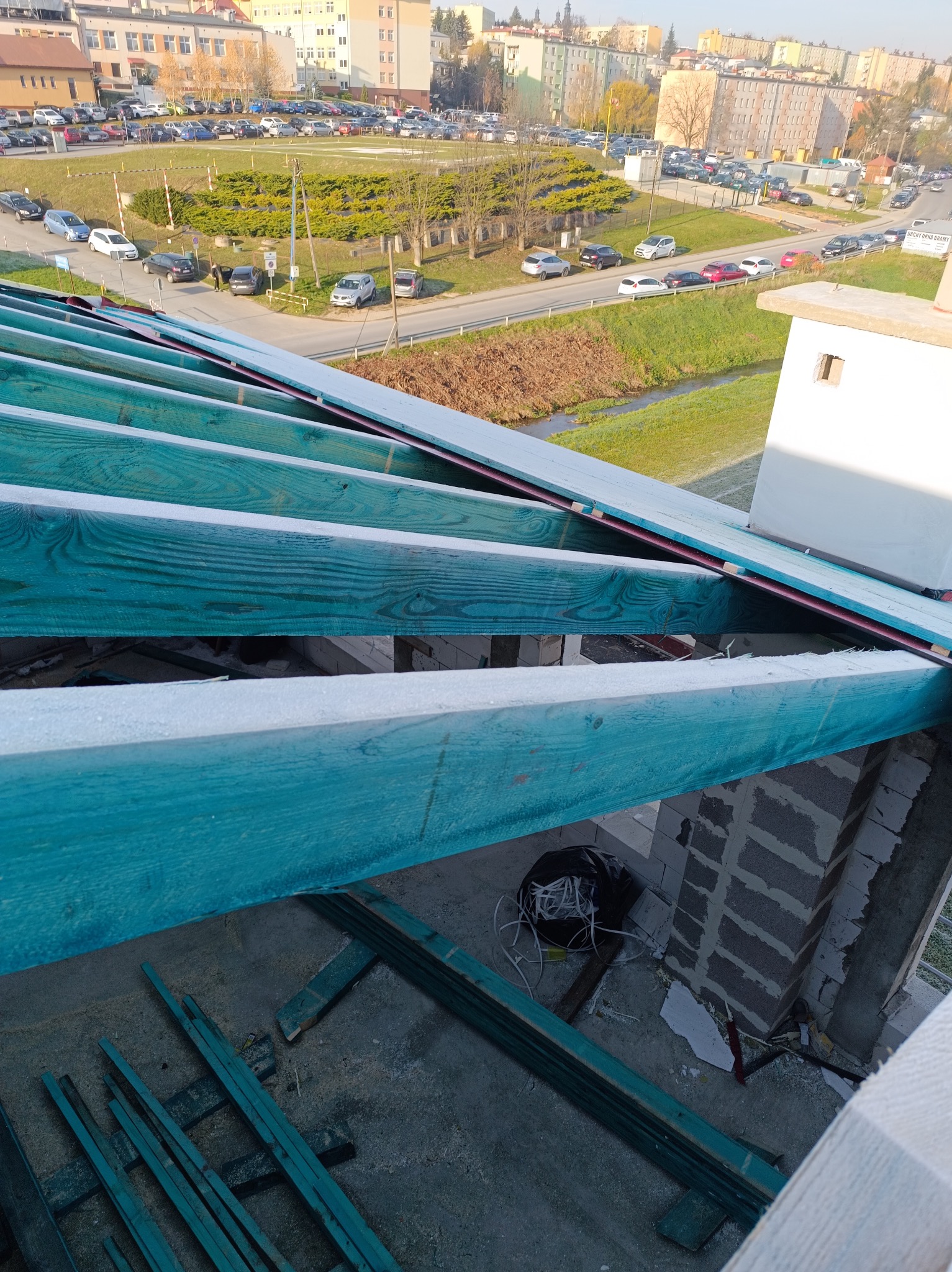 Wiązary dachowe zaimpregnowane na turkusowo, widok z góry na budowę, w tle zabudowa miejska i parking. Elementy więźby dachowej częściowo poza kadrem.