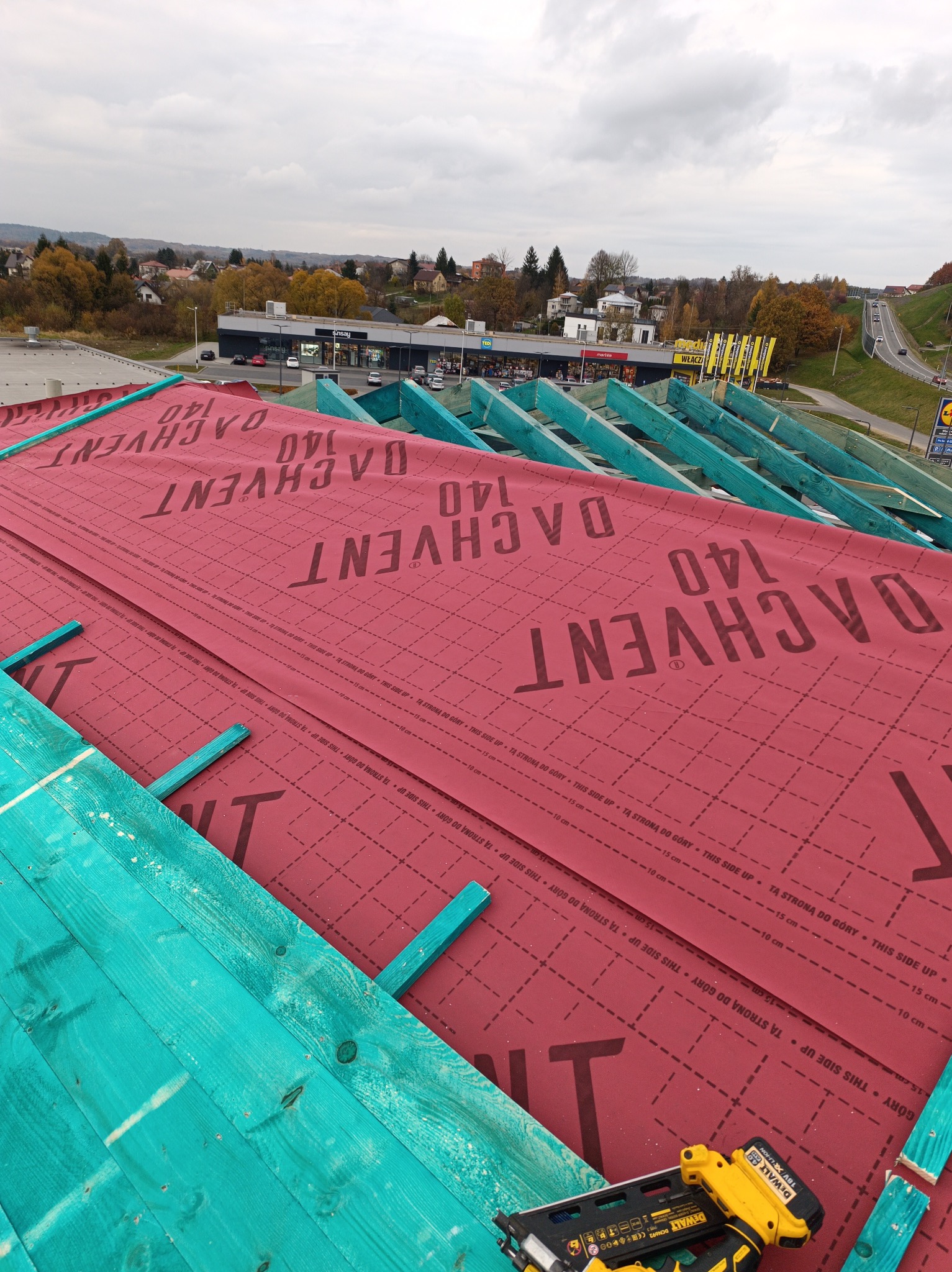 Montaż membrany dachowej Dachvent 140 na drewnianej więźbie w Tyrawie Wołoskiej. Widok z góry na częściowo pokryty dach i narzędzie pracy.