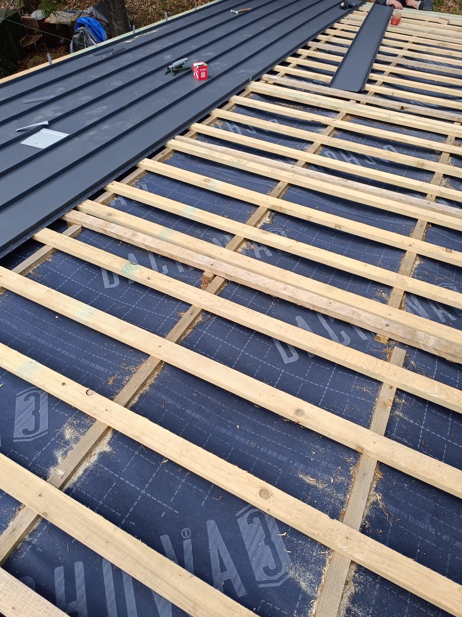 Fragment dachu w trakcie krycia: szara blacha trapezowa ułożona częściowo, widoczna drewniana konstrukcja łat i kontrłat na membranie dachowej z nadrukiem.