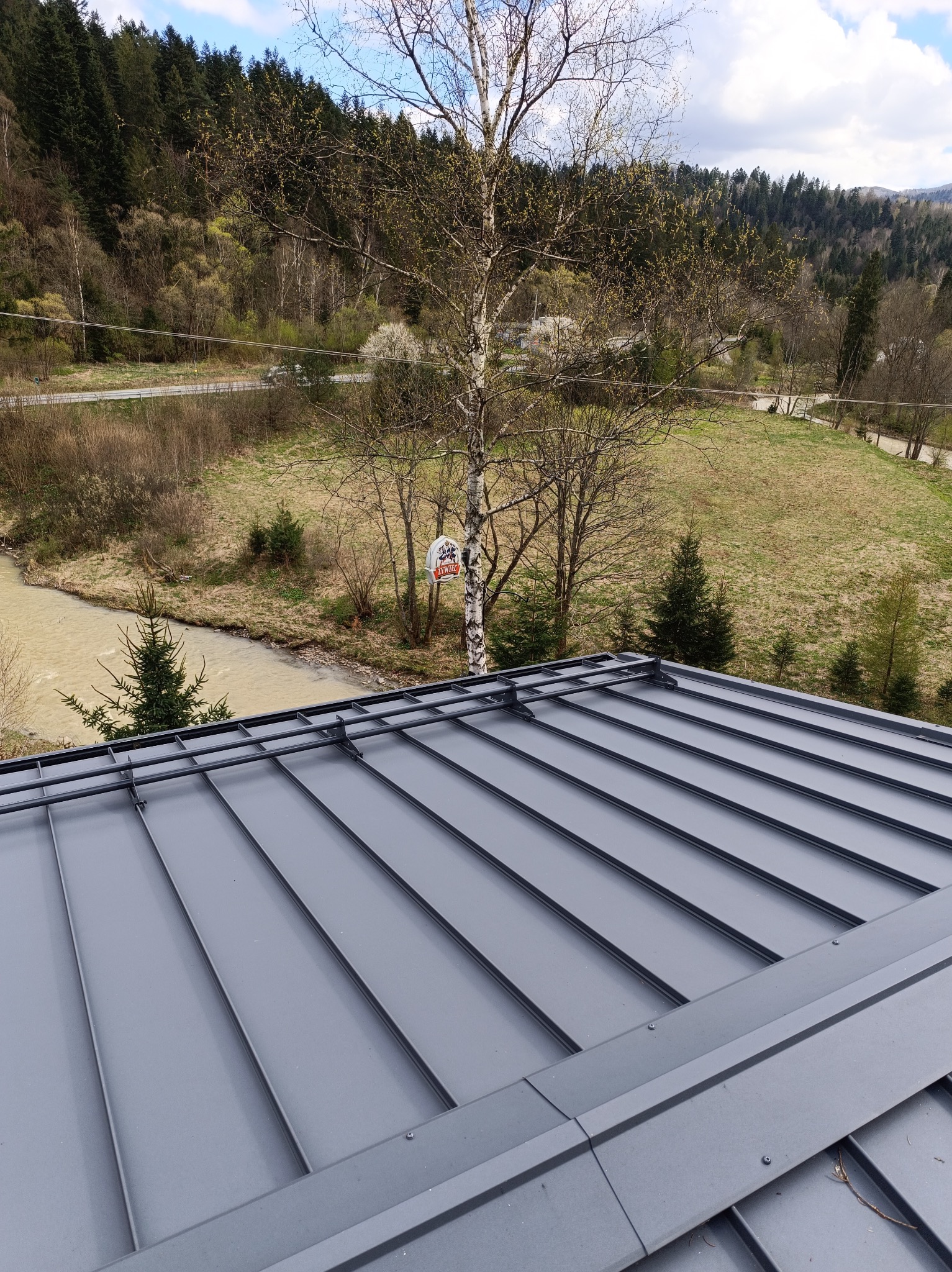 Szary dach panelowy na tle górskiego krajobrazu z rzeką i brzozą z reklamą. Widok z góry na nową, metalową konstrukcję dachu.