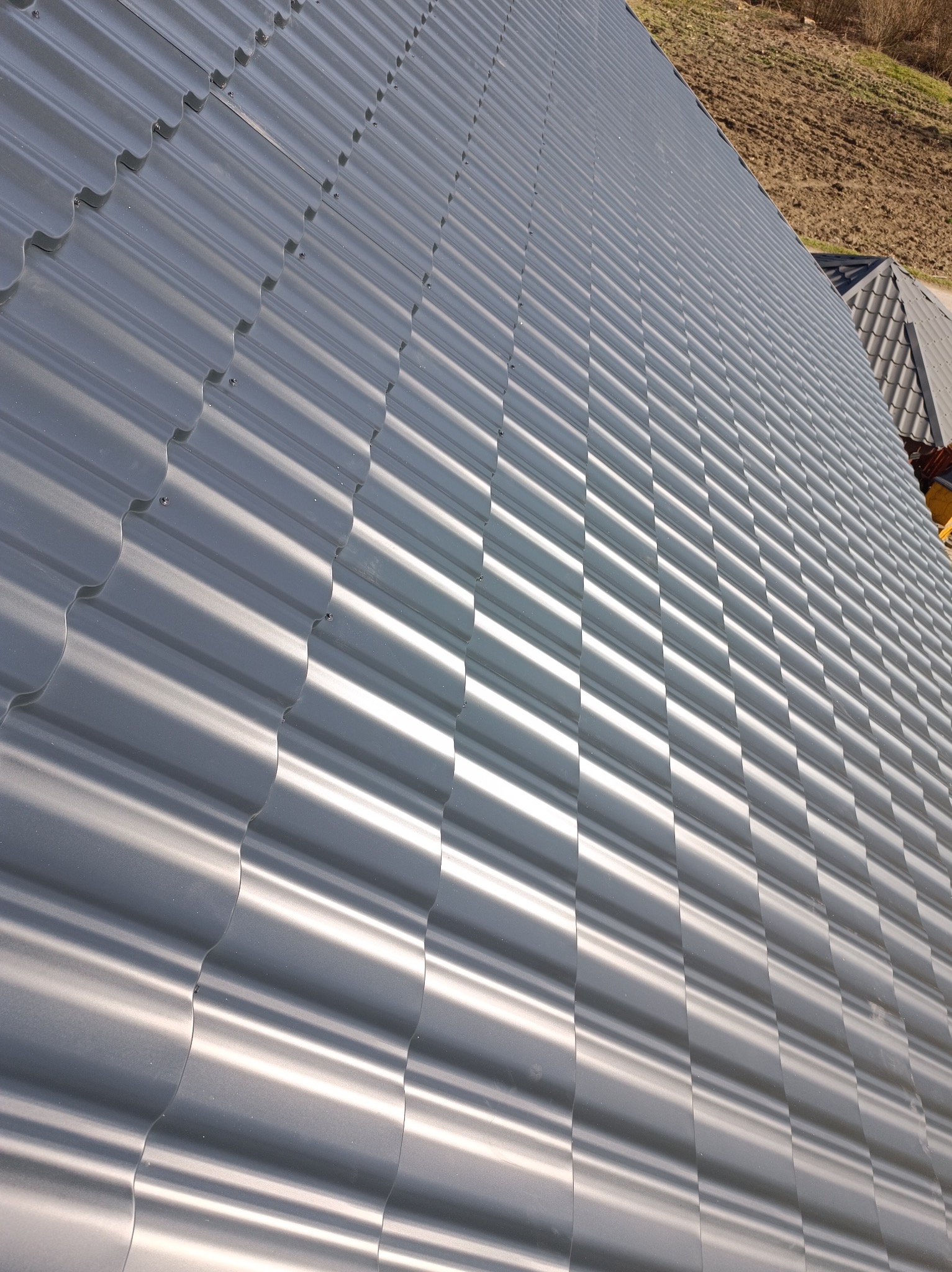 Szary dach z blachy trapezowej w ukośnym ujęciu, widoczne mocowania. W tle fragment pola i dachu sąsiedniego budynku.