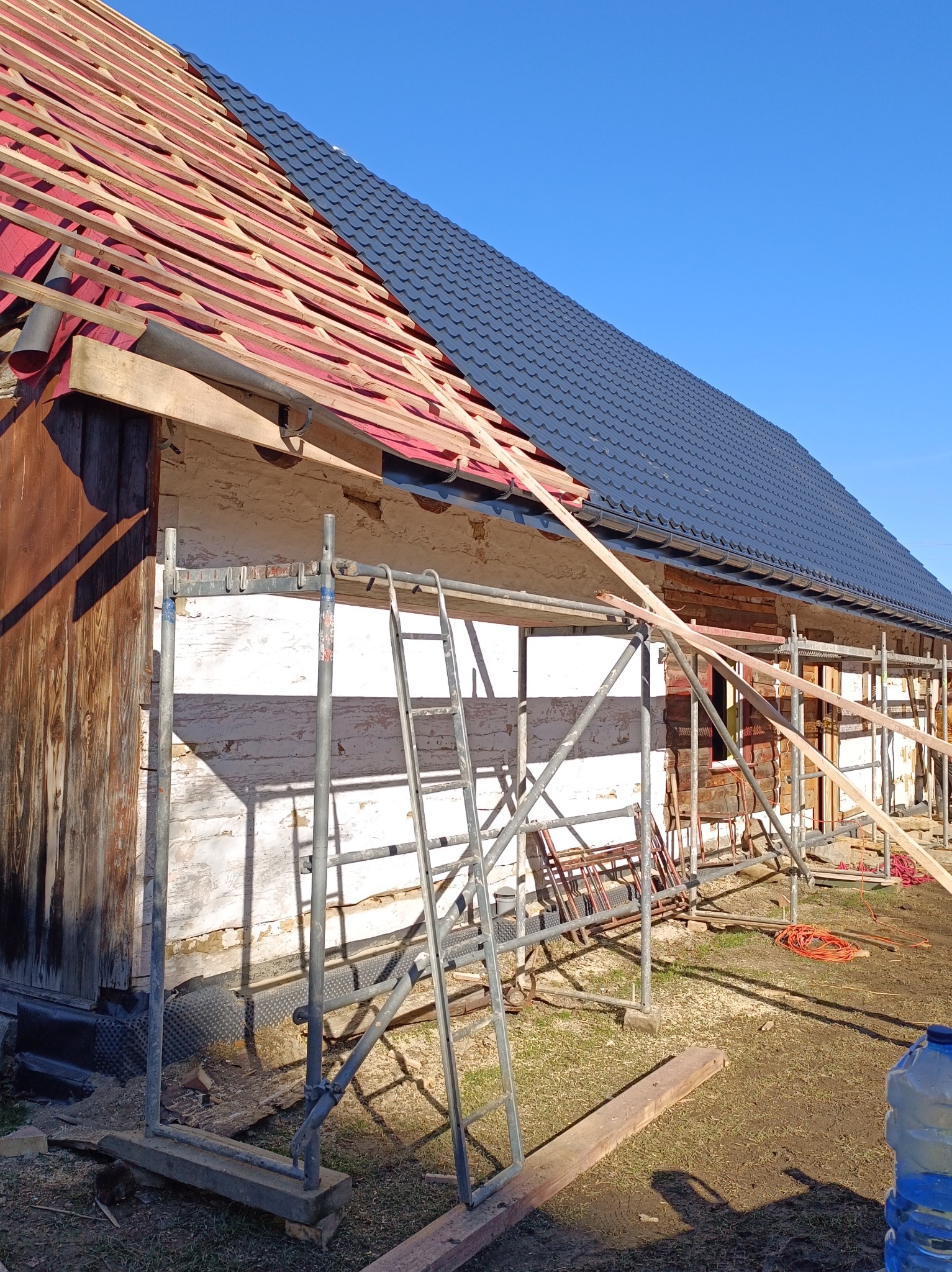 Częściowo pokryty, ciemny dach z blachy trapezowej na drewnianej konstrukcji domu. Widoczne rusztowania i elementy konstrukcyjne dachu. Prace dekarskie w toku.