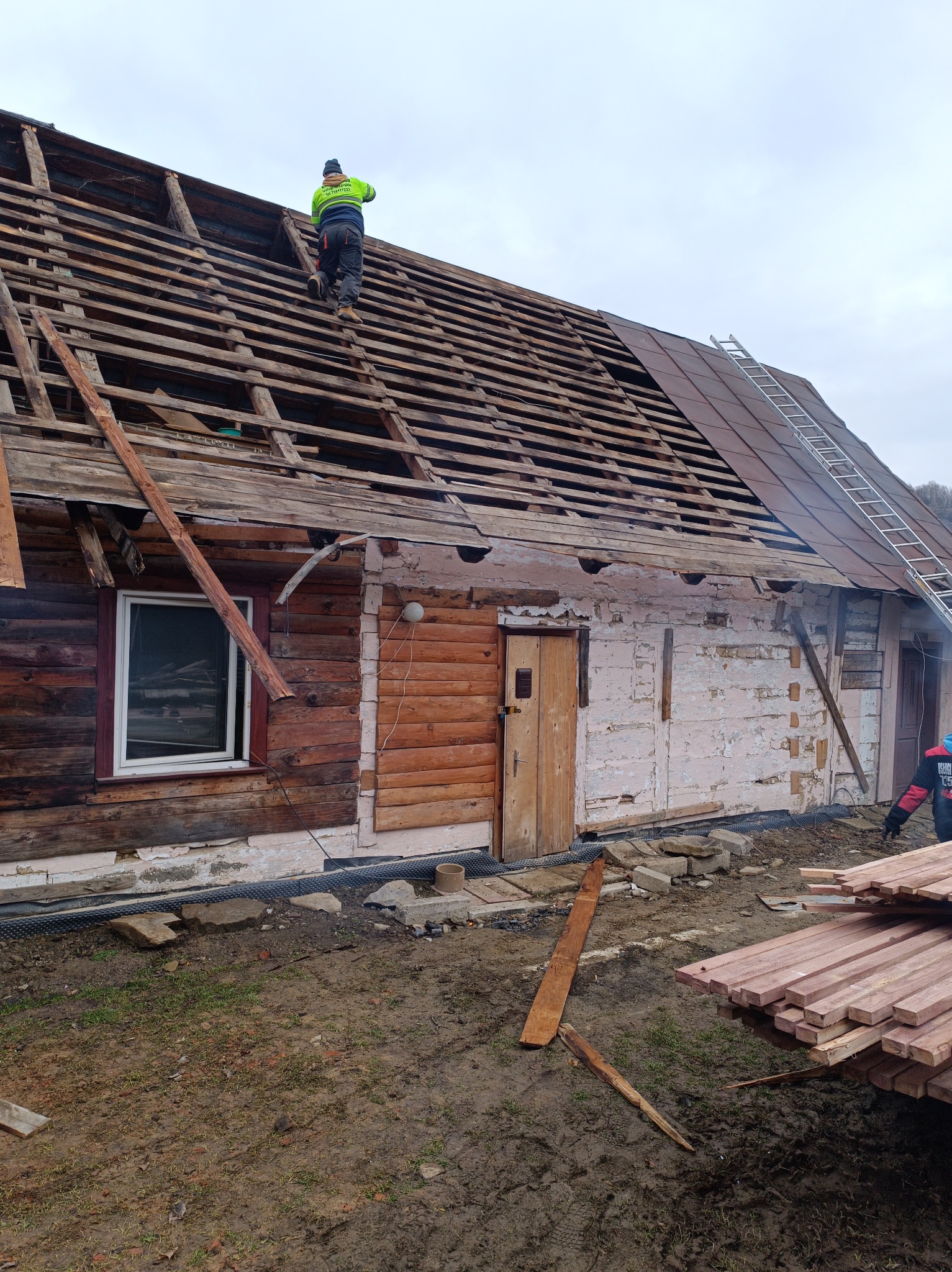 Demontaż starego dachu drewnianego domu. Widoczna więźba dachowa i częściowo zdjęte pokrycie. Na dachu pracuje dekarz w jaskrawej kamizelce. Przygotowane nowe deski.