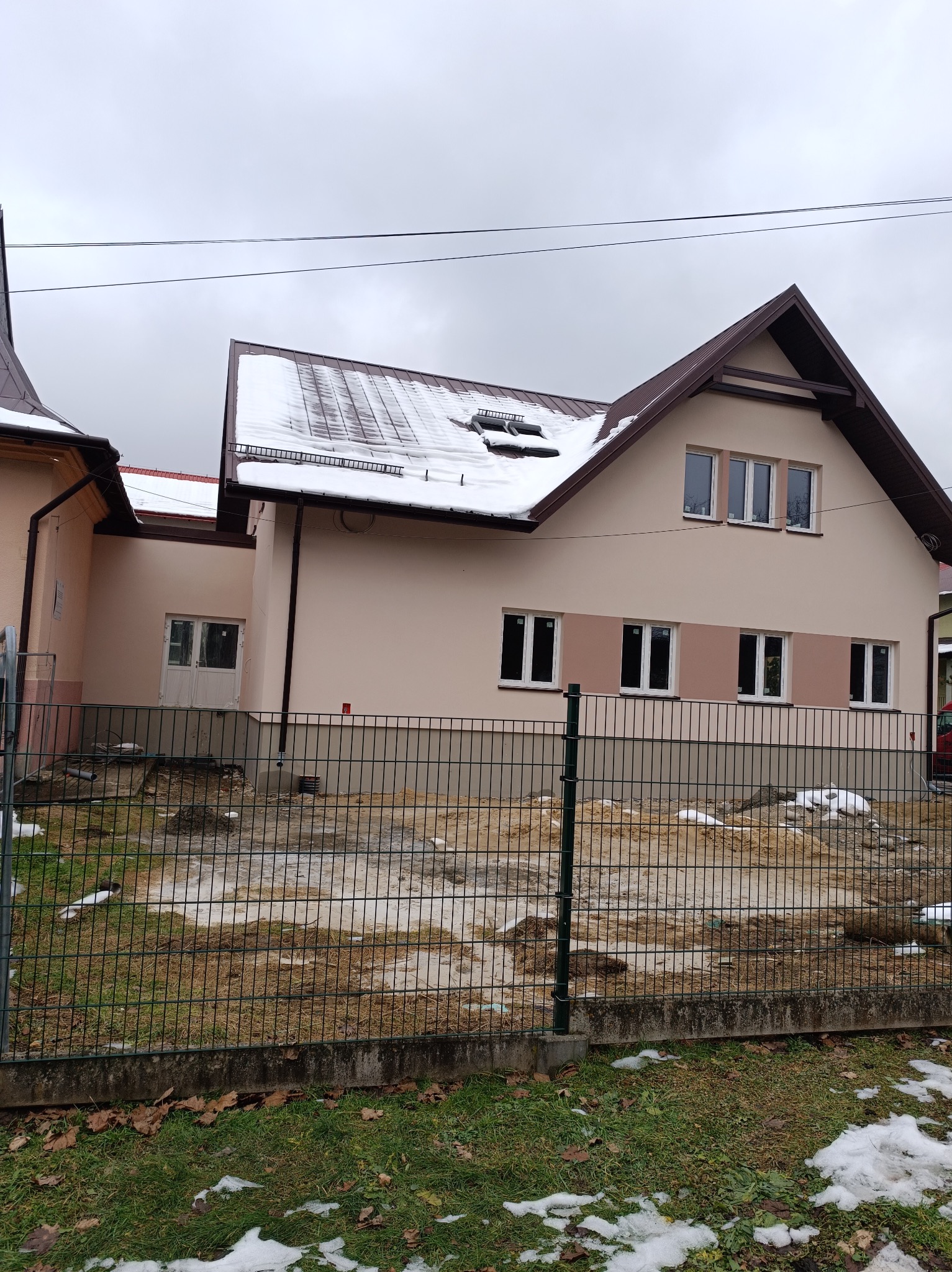 Dom z brązowym dachem pokrytym śniegiem, widoczny zza ogrodzenia. Elewacja w odcieniach beżu, z brązowymi obramowaniami okien i dachu. Pochmurne niebo w tle.