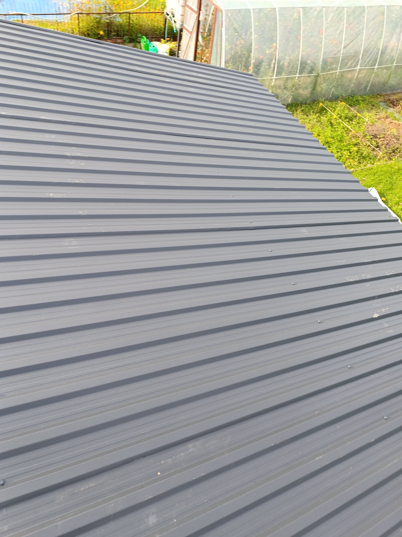 Szary dach z blachy trapezowej, widok z góry. W tle zieleń i fragment szklarni.