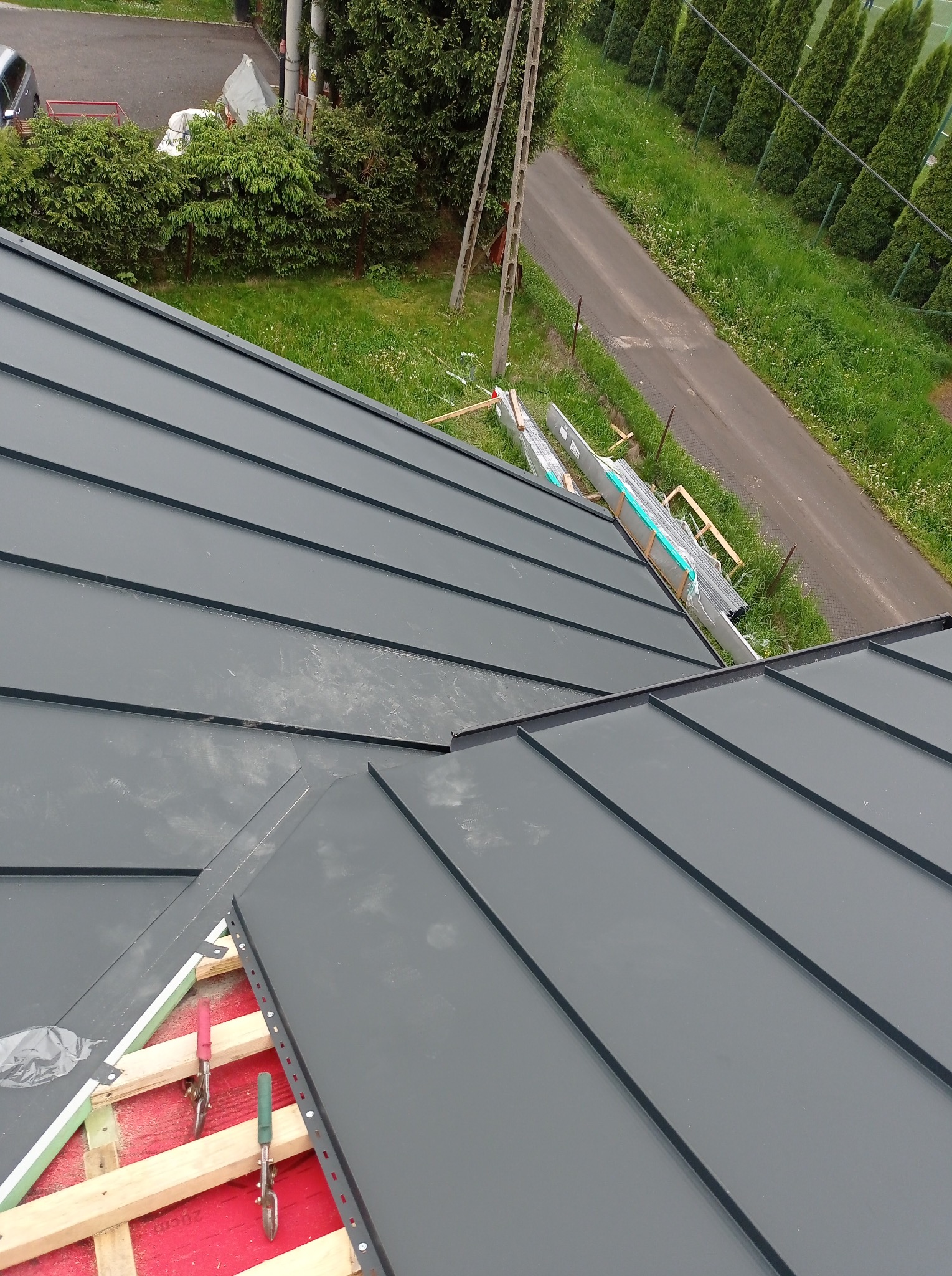 Montaż blachy na dachu. Widoczne narzędzia dekarskie, elementy konstrukcji i blacha w kolorze grafitowym. W tle zieleń i droga.