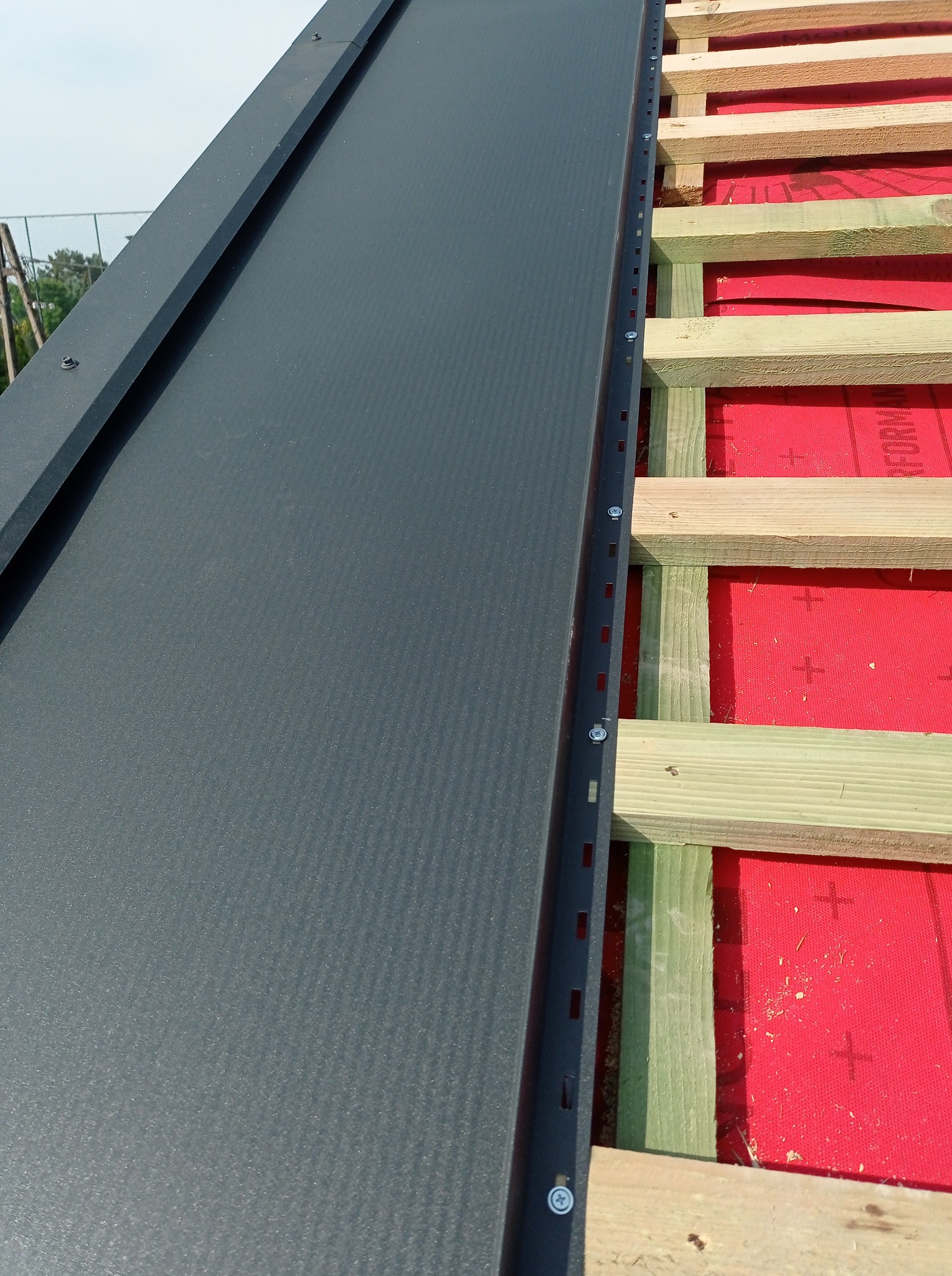 Fragment dachu w trakcie montażu: szara blacha na rąbek stojący, listwa montażowa z widocznymi śrubami, drewniana konstrukcja i czerwona membrana dachowa.