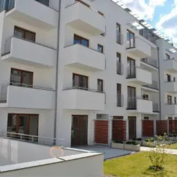 Nowoczesny budynek wielorodzinny z białymi elewacjami, balkonami z metalowymi balustradami i brązowymi oknami. Widoczne zadbane trawniki i elementy małej architektury.