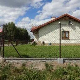 Ciemna siatka ogrodzeniowa na betonowej podmurówce, zabezpieczająca posesję z domem jednorodzinnym w tle, w słoneczny dzień.