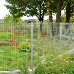 Panelowe ogrodzenie posesji z widocznymi słupkami i podmurówką, na tle zieleni i zabudowań mieszkalnych. Widok z zewnątrz, w dzień.