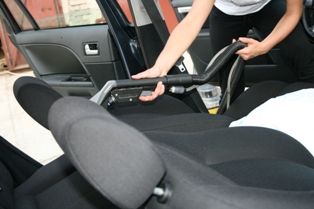 Czyszczenie wnętrza samochodu odkurzaczem z wąską końcówką, widoczne fotele z zagłówkami, pasy bezpieczeństwa i fragment otwartych drzwi.