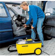 Kobieta odkurza wnętrze niebieskiego samochodu przy użyciu żółtego odkurzacza piorącego Karcher. Widoczny otwarty bok samochodu i fragment deski rozdzielczej.