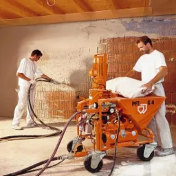 Dwóch mężczyzn w białych koszulkach nakłada tynk maszynowy na ścianę z cegły, używając pomarańczowej maszyny PFT G4 z workiem zaprawy.