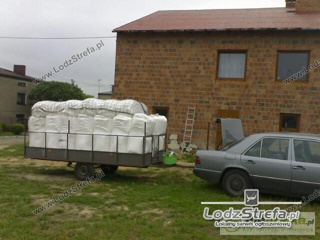 Przyczepa z workami granulatu styropianowego na tle ceglanego domu z oknami, obok zaparkowany szary samochód, widoczna drabina oparta o ścianę.