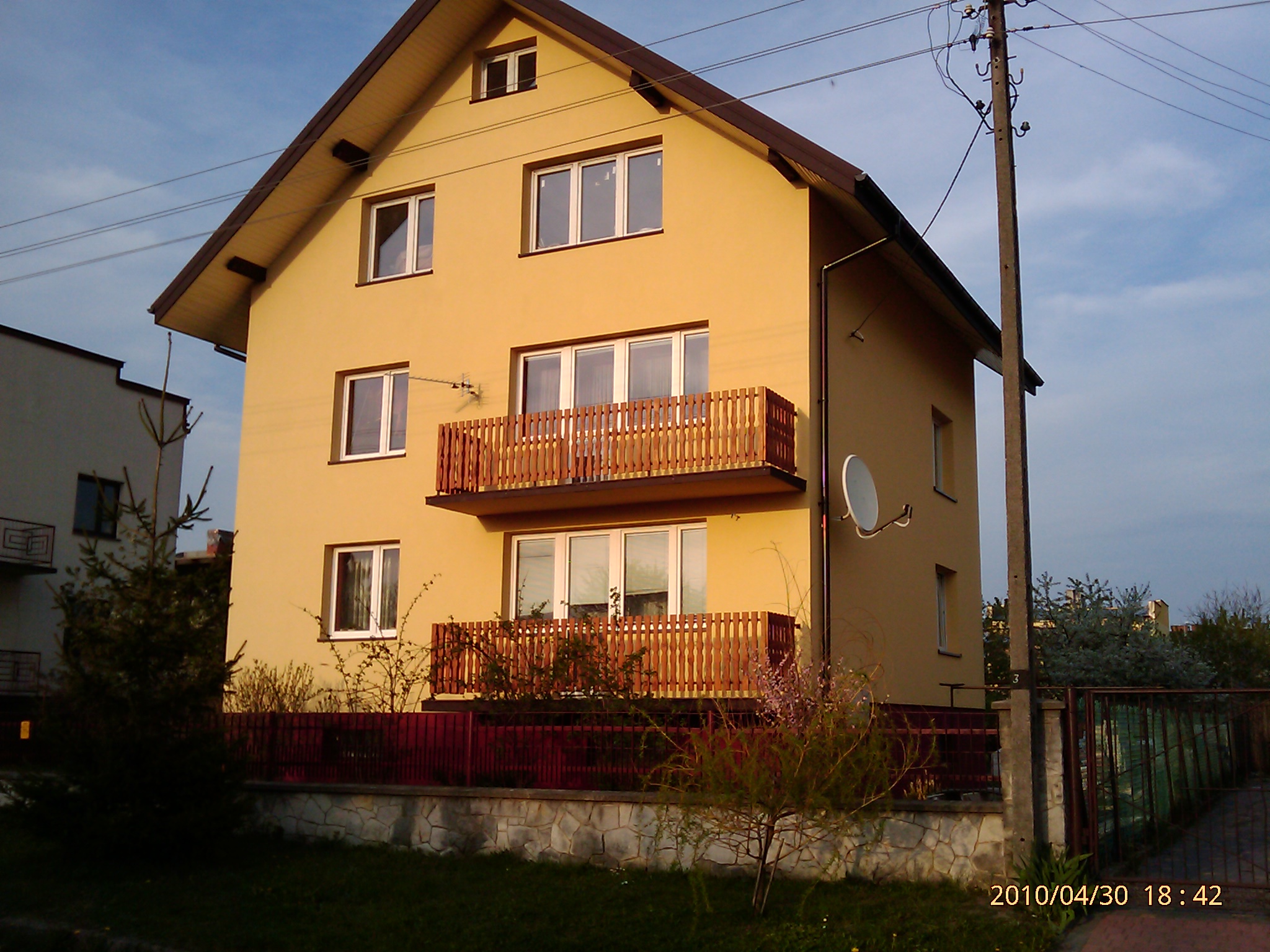 Żółty dom jednorodzinny z dwoma balkonami z drewnianymi balustradami, widoczny z zewnątrz, z czerwoną metalową siatką ogrodzeniową na kamiennej podmurówce, antena satelitarna na ścianie bocznej...