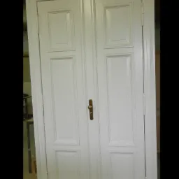 Odrestaurowane, białe, dwuskrzydłowe drzwi z dekoracyjnymi panelami i mosiężną klamką, ukazane w zbliżeniu.