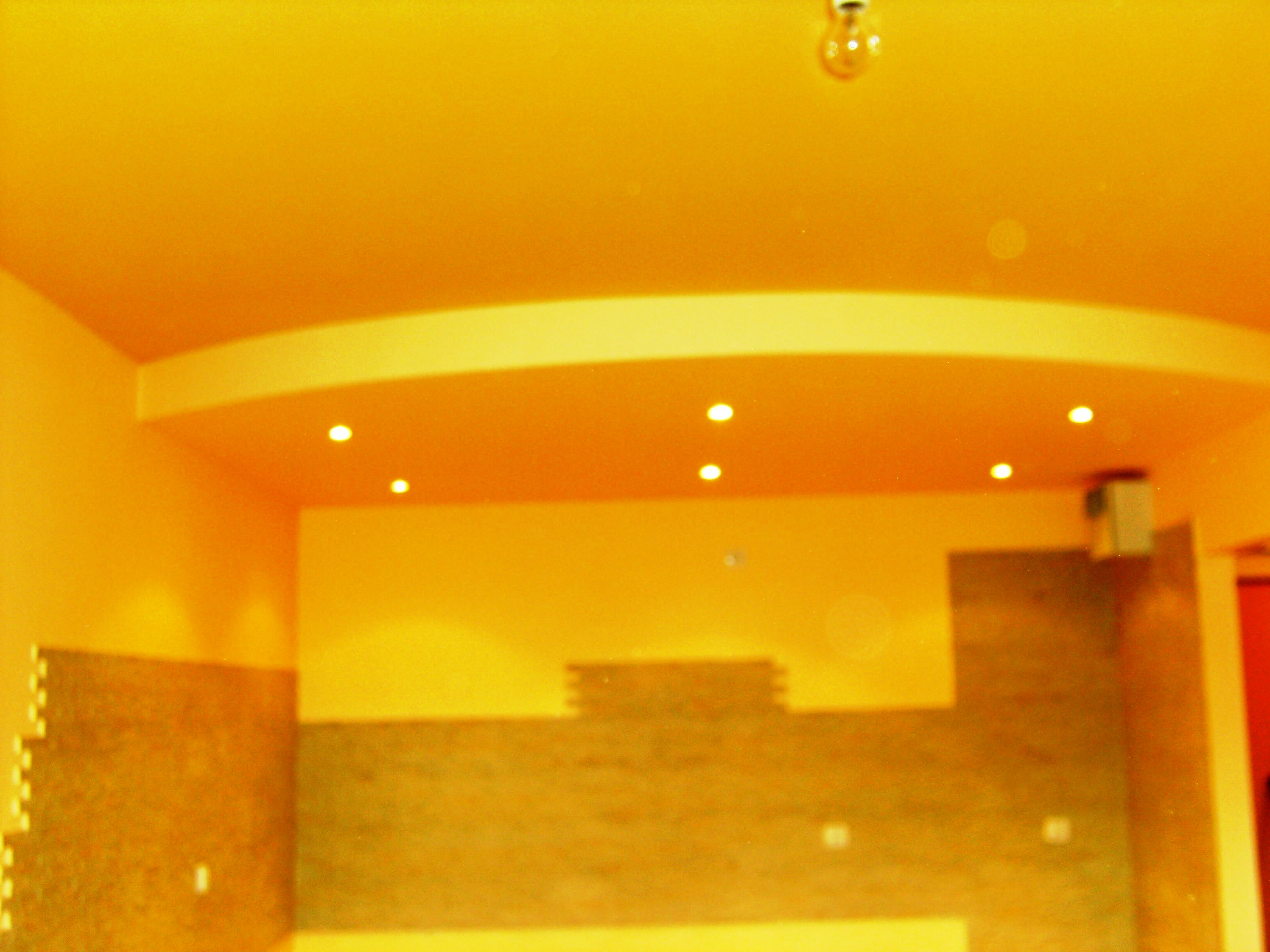 Żółte wnętrze pokoju w trakcie remontu, widoczne fragmenty ścian z różnym wykończeniem i oświetleniem punktowym.