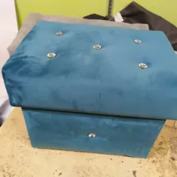 Turkusowy, welurowy puf z dekoracyjnymi guzikami na blacie i pojedynczym guzikiem na froncie dolnej części, stojący na stole w warsztacie.