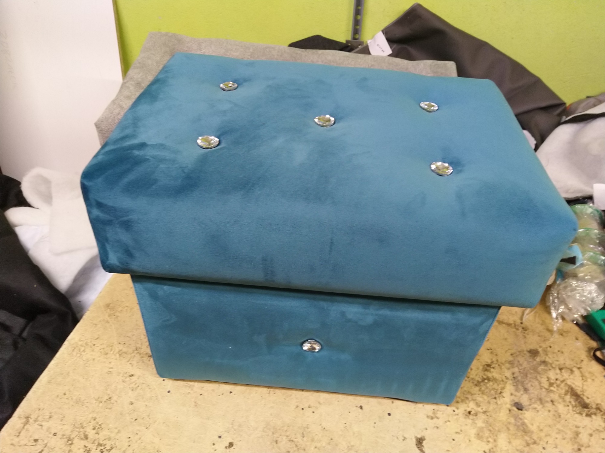 Turkusowy, welurowy puf z dekoracyjnymi guzikami na blacie i pojedynczym guzikiem na froncie dolnej części, stojący na stole w warsztacie.