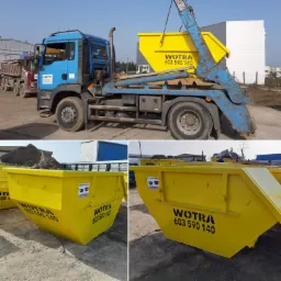 Żółty kontener na gruz podnoszony przez niebieską ciężarówkę z hydraulicznym ramieniem, widoczne inne kontenery i ciężarówki w tle.